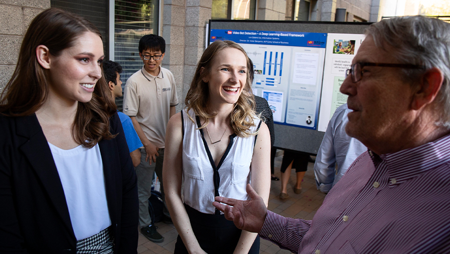 Two students speaking with an adult with their thesis in the background